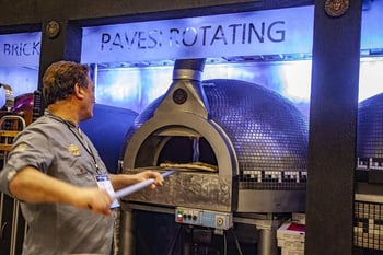 Man in grey chef's coat uses paddle to load pizza into Fiero Pavesi rotating pizza oven. 