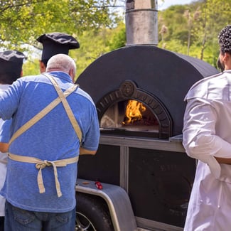 Man in blue shirt and tan apron with back to camera, look in black pizza oven on trailer