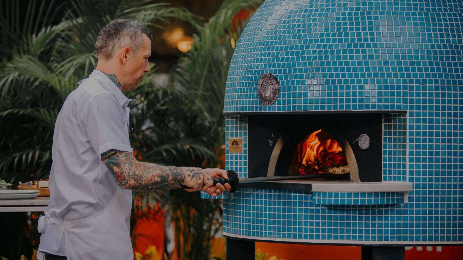Chef in white apron and tatoos is outside inserting a pizza into a blue mosaic tile Fiero wood-fired pizza oven.