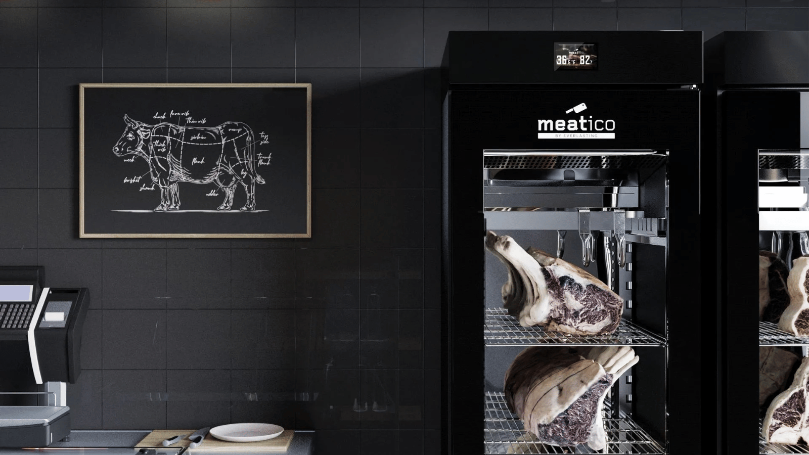Meatico dry age meat vertical cabinet stands against a black wall. To the left of the cabinet hangs a picture of a diagram of a cow with the different cuts of meat indicated on it. 