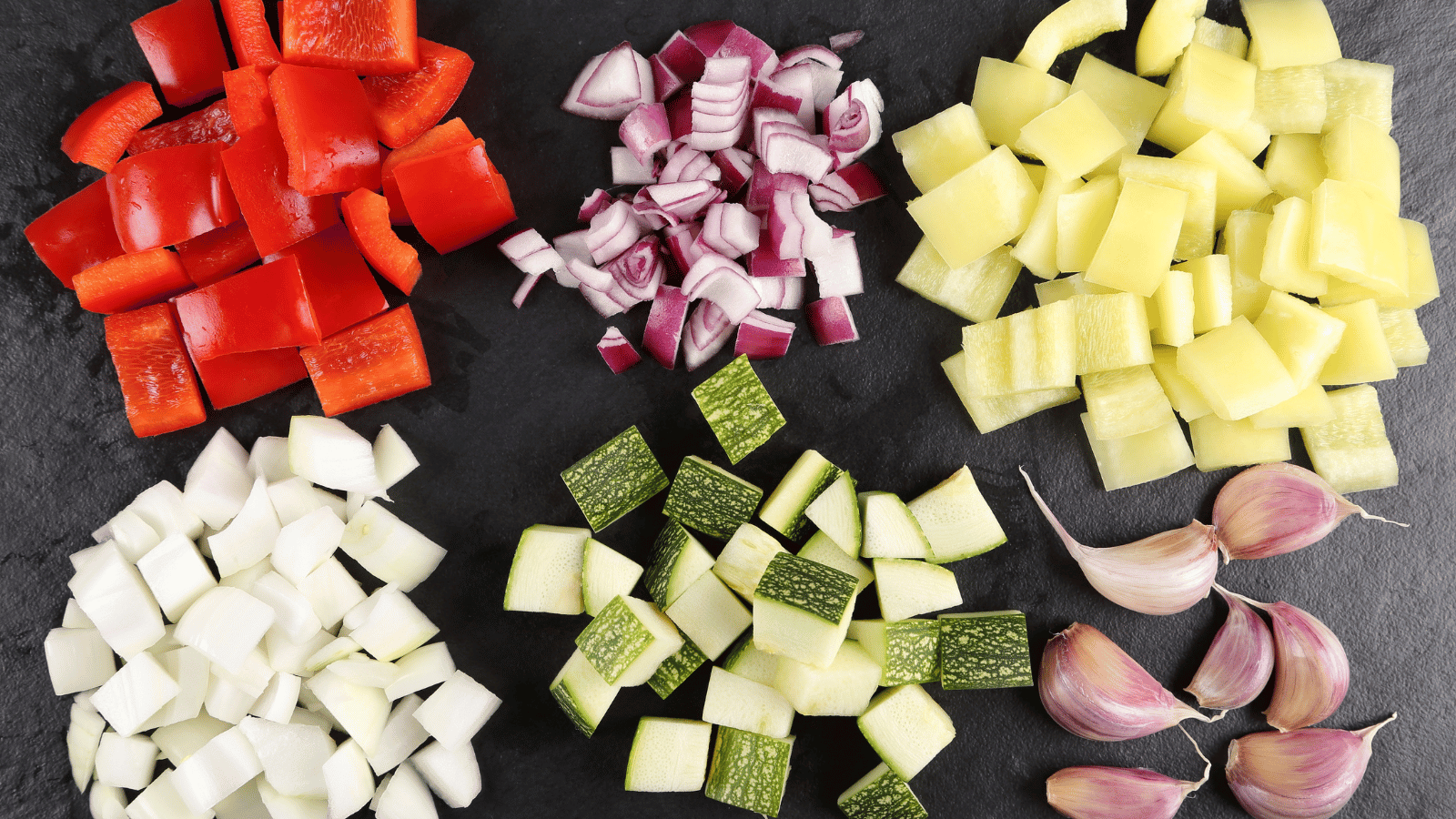 Five sets of diced vegetables lined up on granite board with whole cloves of garlic in bottom right