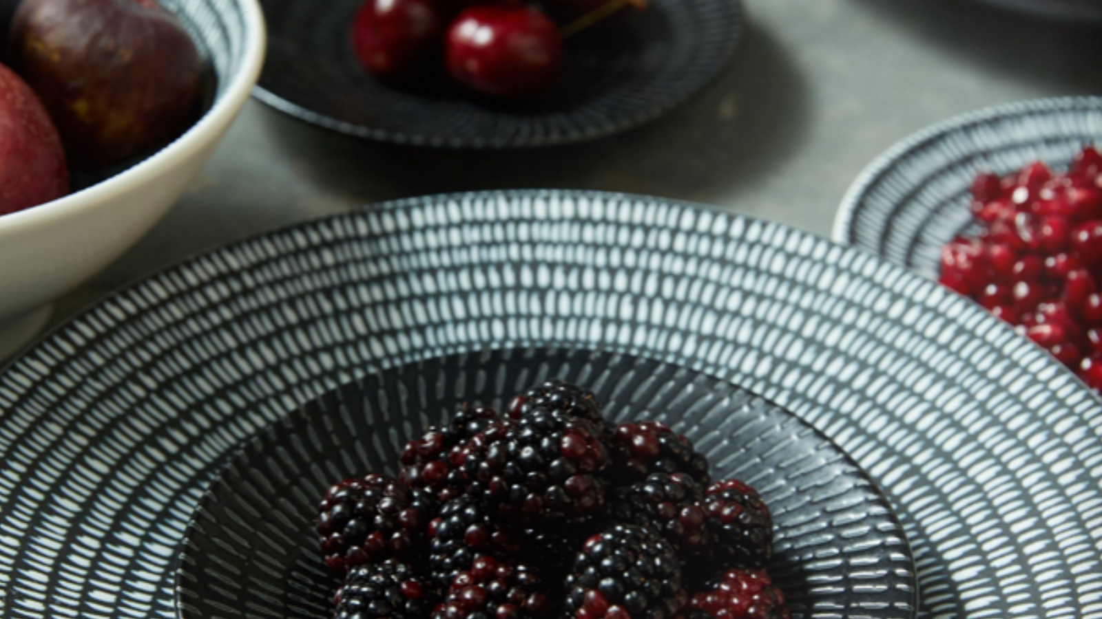A large stone dinner plate with and blue dotted lines in a circle around the entire face is on top of a dinner table with a white linen cloth.. The plate is covered with a large pile of dark black raspberries. Behind the plate is a smaller blue pate with a few fresh, unpitted cherries. Each cherry still has its stem.