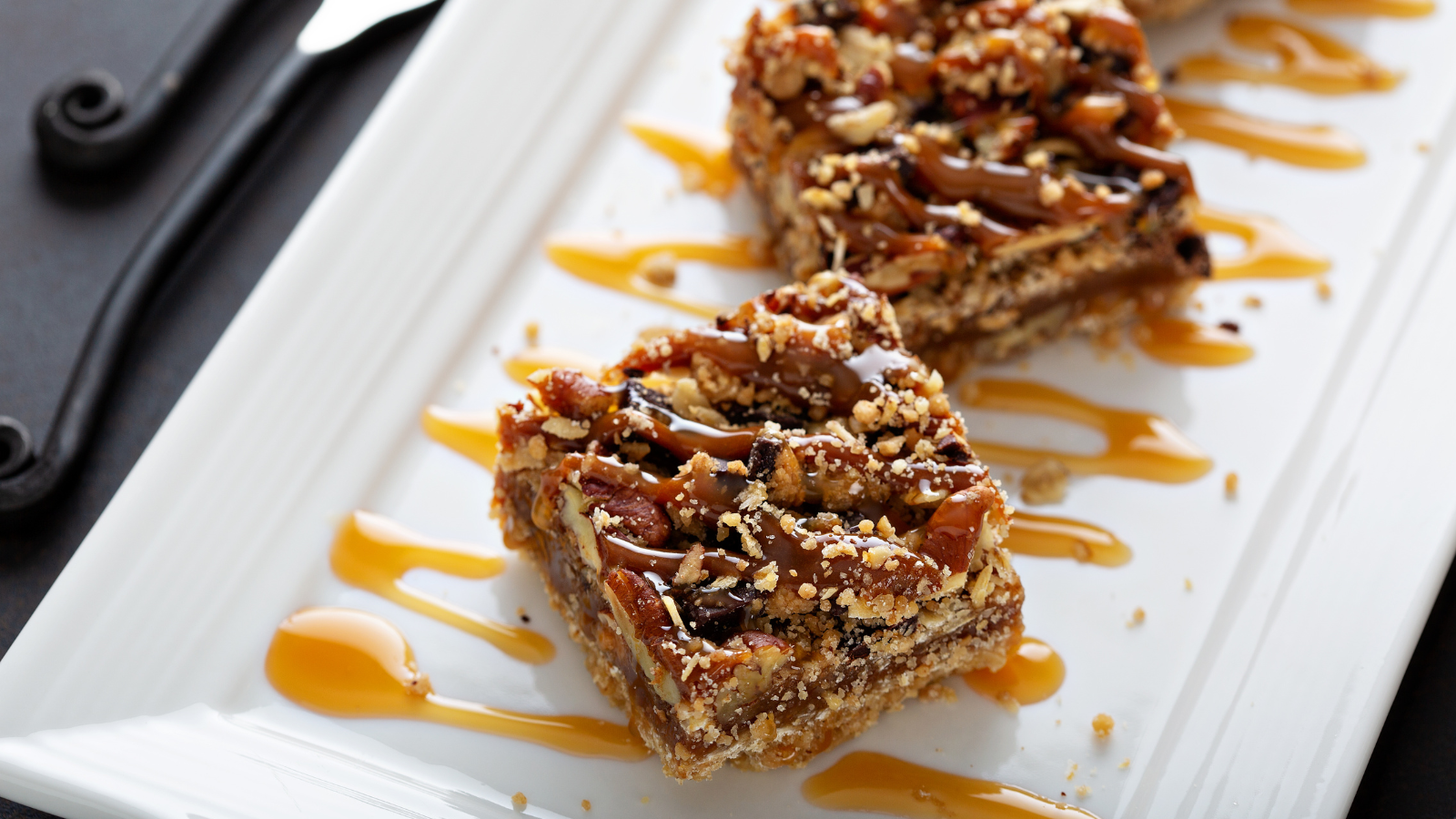 A long white porcelain plate is topped with two square Pecan Sandies that look gooey and sticky. The Pecan Sandies are placed on top of caramel drizzles covering the bottom of the dessert dish.