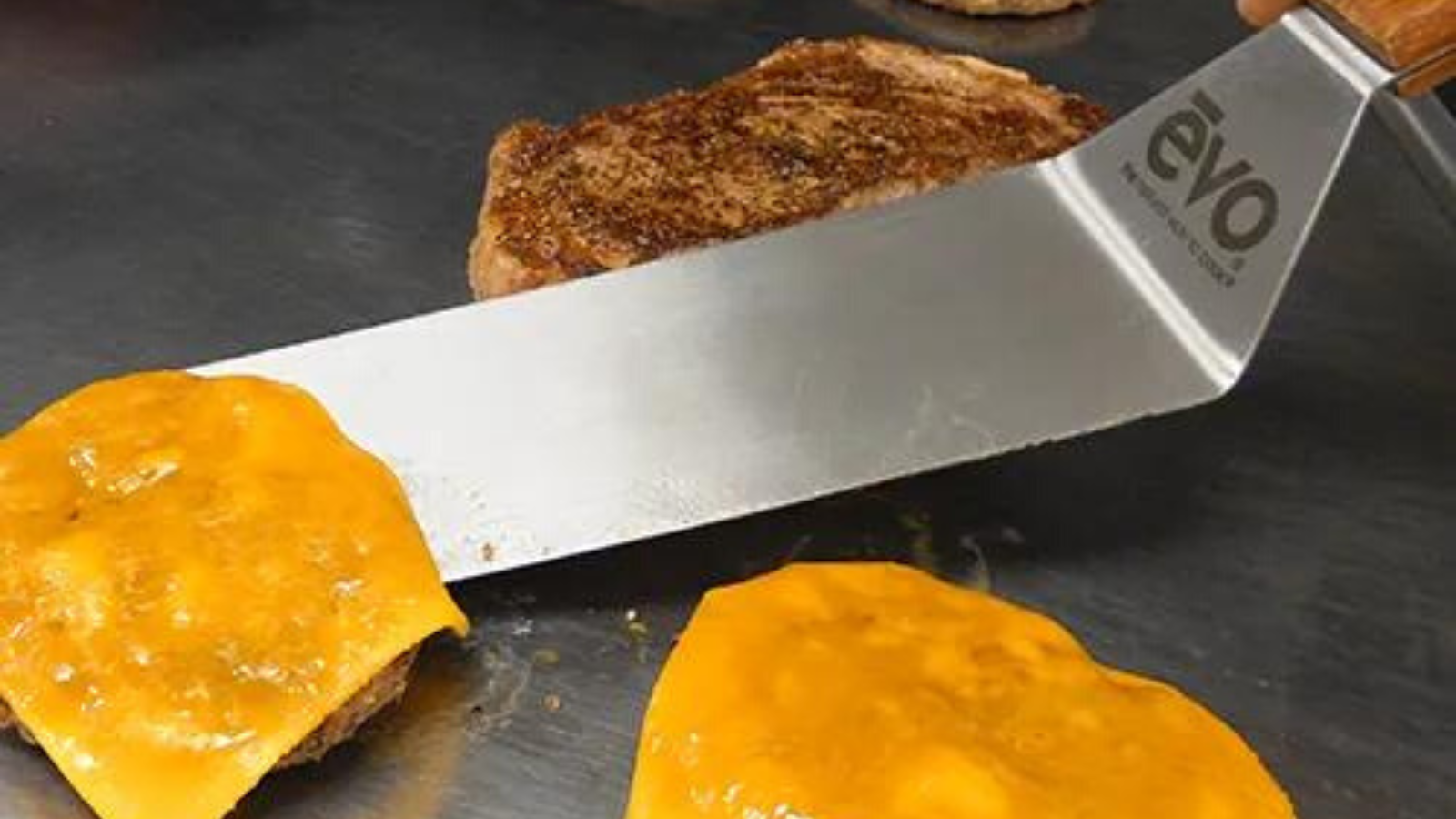 Long spatula with Evo logo on top sliding under one cheeseburger on griddle. Hamburger on griddle to left of spatular and another cheeseburger on griddle to right of spatula.